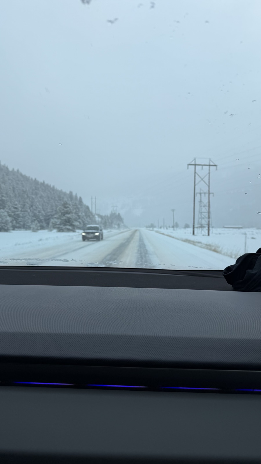 Snow-covered two-lane road ahead through a windshield, frosted evergreens and power lines alongside